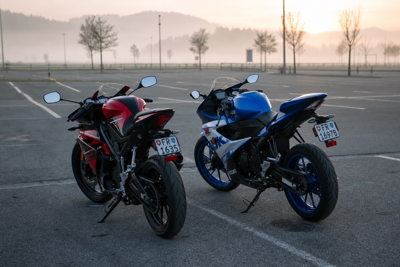 Zwei 125er-Sportmotorräder, eine in Rot und Schwarz, eine in Blau und Weiß, Seite an Seite auf einem leeren Parkplatz in der Morgendämmerung